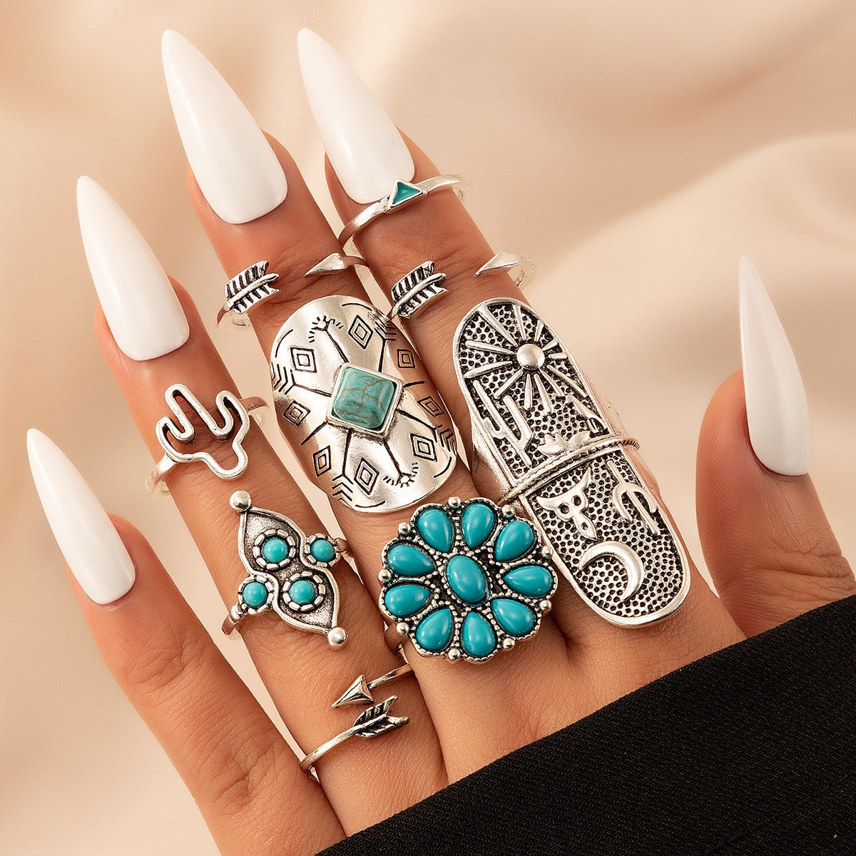 Hand with long white nails wearing a stack of silver boho rings with turquoise stones, arrow, cactus, and sun motifs